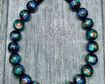 Puede incluir: Un collar hecho de cuentas iridiscentes azules, verdes y doradas con un cierre negro.