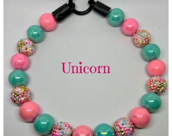 May include: A pink and teal beaded necklace with a black clasp. The beads are decorated with glitter and rhinestones. The necklace has the word "Unicorn" written on it.