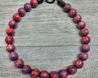 Puede incluir: Un collar para perros con cuentas veteadas rojas y azules. Las cuentas están unidas con un cierre y conectores de metal negro. El collar está diseñado para mascotas.