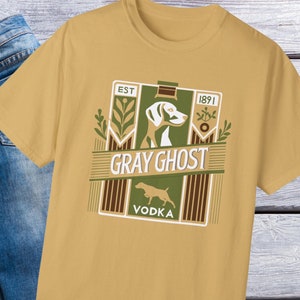 May include: A mustard yellow t-shirt with a vintage-style illustration of a bottle of vodka. The label reads "Gray Ghost Vodka" with the text "Est. 1891" and a stylized image of a dog.