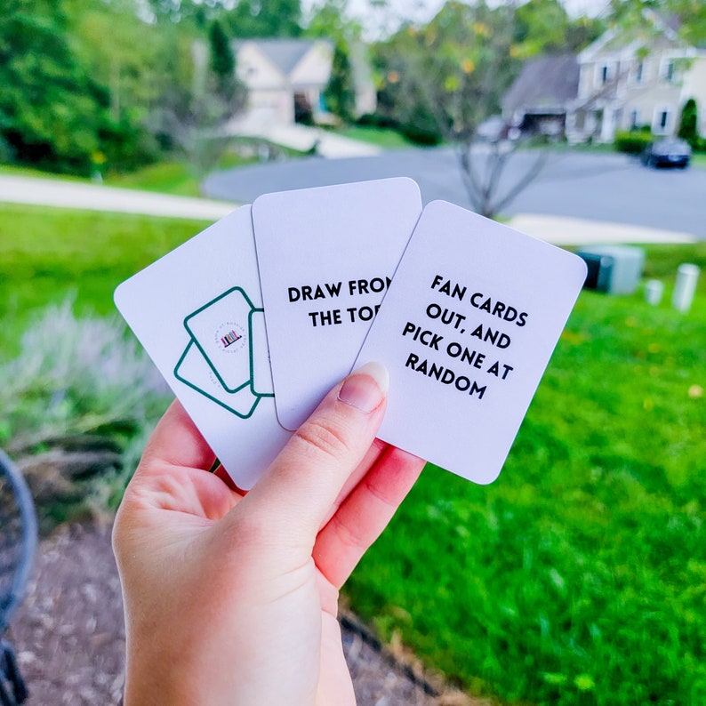 May include: Three white cards with black text. The top card says "Fan cards out, and pick one at random". The middle card says "Draw from the top". The bottom card has a green outline of a stack of books.