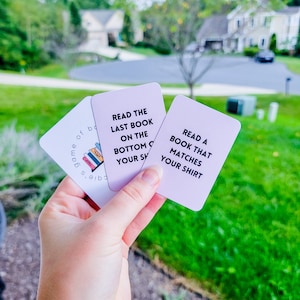 May include: Three pink cards with white text. The top card says "Read the last book on the bottom of your shelf". The middle card says "Read a book that matches your shirt". The bottom card says "Game of Books".