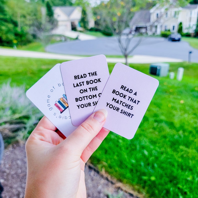 May include: Three pink cards with white text. The top card says "Read the last book on the bottom of your shelf". The middle card says "Read a book that matches your shirt". The bottom card says "Game of Books".