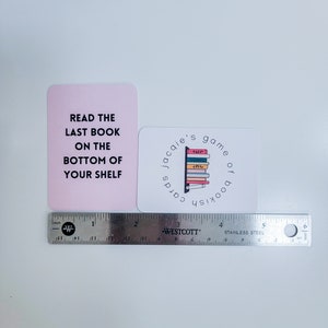 May include: Two pink and white cards with text that reads "Read the last book on the bottom of your shelf" and "Jacquie's game of bookish cards". The second card has a graphic of a bookshelf with colourful books.