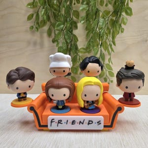 Könnte beinhalten: Sechs Figuren der Charaktere aus der Serie Friends, jede mit einer einzigartigen Frisur und Kleidung. Sie sind um ein orangefarbenes Sofa angeordnet, auf dem das Wort "FRIENDS" steht. Der Hintergrund umfasst Grün und eine hellfarbene Holzoberfläche.