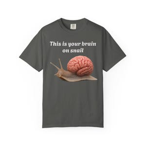 Op de afbeelding: Een antracietgrijs T-shirt met korte mouwen en een afbeelding van een slak met een menselijk brein als huisje. Boven de slak staat de witte tekst, This is your brain on snail. Het shirt heeft een ronde hals en een standaard pasvorm.