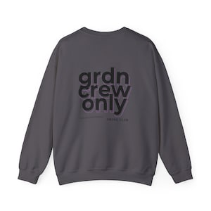 Peut inclure: Un sweat-shirt gris anthracite avec le texte "grdn crew only" en noir avec un contour violet. Le texte "SMOKE.CLUB" est imprimé en noir sous le texte principal.