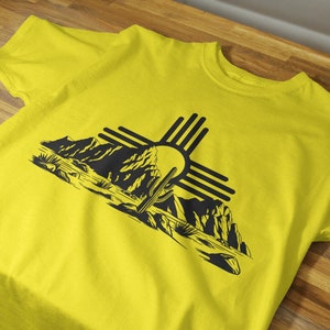 May include: A yellow t-shirt with a black graphic print of a desert landscape with a cactus and the Zia symbol.