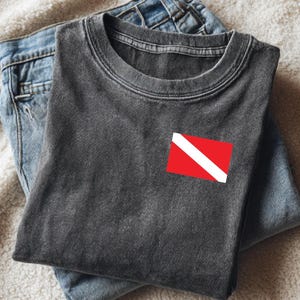 May include: A dark gray t-shirt with a scuba diving flag patch on the chest, folded on top of a pair of blue jeans. The scuba flag is red with a white diagonal stripe. The shirt and jeans are on a white, textured surface.
