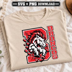 Puede incluir: Una camiseta beige de manga corta con un gráfico de un caballo blanco con crin roja irrumpiendo a través de una gran letra B roja. La palabra BRONCOS está impresa verticalmente en letras rojas junto al caballo. El diseño tiene una textura desgastada.