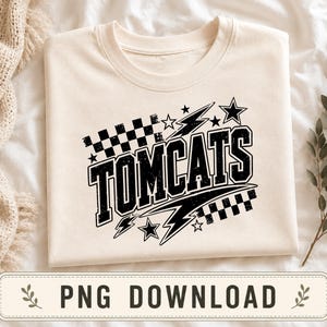 Może przedstawiać: Kremowy t-shirt z czarnym nadrukiem graficznym przedstawiającym słowo TOMCATS pogrubioną, postarzaną czcionką typu varsity. Wzór zawiera motywy szachownicy, błyskawic i gwiazd ułożone w dynamiczny, sportowy układ graficzny na środku koszulki.