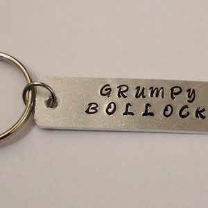 Könnte beinhalten: Silberner Schlüsselanhänger mit einem silbernen Ring und einem rechteckigen Anhänger mit der Aufschrift "GRUMPY BOLLOCKS" in schwarzer Schrift.
