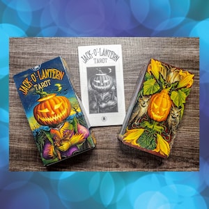 Puede incluir: Tres mazos de cartas del tarot Jack-O'-Lantern y un folleto se muestran sobre una superficie de madera. Las barajas presentan ilustraciones vibrantes de calabazas, búhos y follaje. El texto "Jack-O'-Lantern Tarot" es visible en las barajas y el folleto.