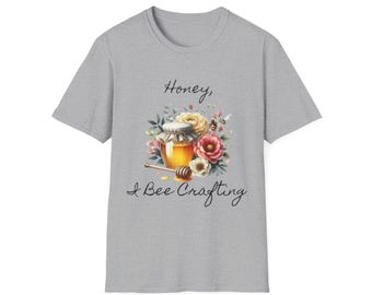 May include: A gray t-shirt featuring a watercolor design of a honey jar, flowers, and a bee. The text "Honey, I Bee Crafting" is written above and below the image. The shirt is made of a soft material.