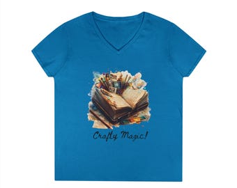 May include: A bright blue V-neck t-shirt featuring a central graphic of an open book surrounded by various art supplies and watercolor splashes. Below the illustration, the text Crafty Magic! is printed in a dark, cursive script font.