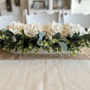 May include: A white ceramic planter filled with white hydrangea flowers and green eucalyptus greenery. The planter is sitting on a burlap runner on a white tablecloth.