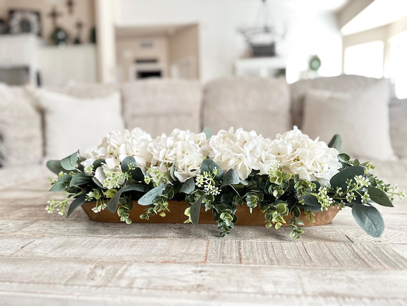 May include: A wooden trough centerpiece filled with white hydrangeas and green eucalyptus leaves.