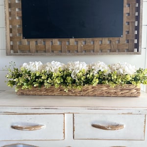May include: A white wicker basket filled with white hydrangea flowers and green foliage sits on a white distressed wooden table with two drawers.