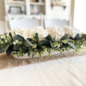 May include: A white rectangular planter filled with white hydrangeas and green eucalyptus leaves. The planter is sitting on a brown burlap runner on a white table.