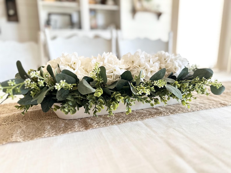 May include: A white rectangular planter filled with white hydrangeas and green eucalyptus leaves. The planter is sitting on a brown burlap runner on a white table.