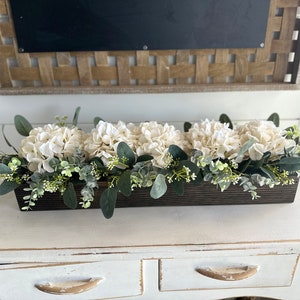 May include: A wooden box planter filled with white hydrangeas and green eucalyptus leaves. The planter is sitting on a white painted dresser.