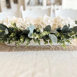 May include: A white ceramic trough planter filled with white hydrangeas and green eucalyptus greenery. The planter is sitting on a burlap runner on a white tablecloth.