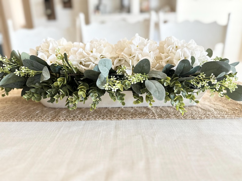 May include: A white ceramic trough planter filled with white hydrangeas and green eucalyptus greenery. The planter is sitting on a burlap runner on a white tablecloth.