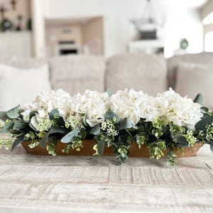 May include: A wooden trough centerpiece filled with white hydrangeas and green eucalyptus leaves.