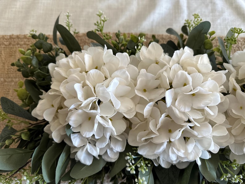 May include: A white hydrangea floral garland with green eucalyptus leaves. The garland is perfect for adding a touch of greenery and elegance to any space.