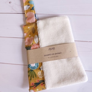 Può includere: Un guanto da bagno bianco con un manico a fantasia floreale giallo senape, rosa, blu e verde. Il guanto da bagno è avvolto con una fascia di carta marrone con testo in italiano.