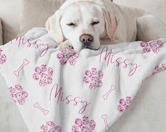May include: A white fleece blanket featuring a repeating pattern of pink floral paw prints and bone outlines, with the name Missy written in a pink cursive script across the fabric. The blanket is draped over a light-coloured sofa.
