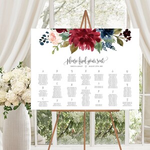 May include: A white wedding seating chart sign on a wooden easel, featuring a floral arrangement of deep red, navy blue, and blush pink flowers at the top. The text reads "please find your seat", "David & Ashley", and "August 27th, 2021".