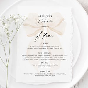 Könnte beinhalten: Eine Menükarte zur Widmung mit einem Aquarell-Schleifendesign, platziert auf einem weißen Teller mit einem Zweig weißer Blumen. Die Speisekarte umfasst Vorspeisen, Hauptgerichte, Desserts und Getränke.