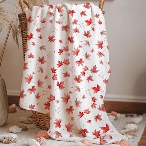 May include: A white blanket with a pattern of red maple leaves is draped over a wicker basket. The blanket has a soft texture and a fringe of pink tassels. The words "forever yours" are printed on the blanket.