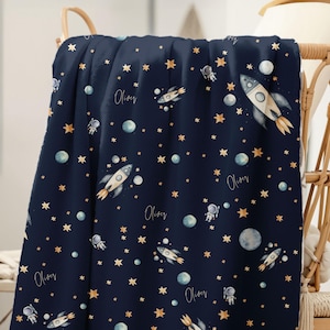 Op de afbeelding: Marineblauwe deken met een ruimtethema. Het ontwerp bevat raketten, astronauten, planeten en sterren in goud, wit en blauw. De naam "Oliver" staat in sierlijke letters over de deken geschreven.