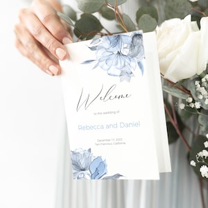 Puede incluir: Un cartel de bienvenida de boda blanco con flores de acuarela azules. El texto dice "Bienvenido a la boda de Rebecca y Daniel, 17 de diciembre de 2023, San Francisco, California."