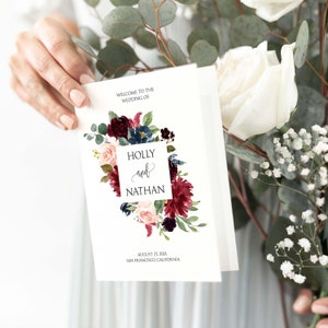 May include: Wedding invitation with a floral design featuring burgundy, blush pink, and navy blue flowers. The text reads "Welcome to the Wedding of Holly and Nathan, August 27, 2021, San Francisco, California."