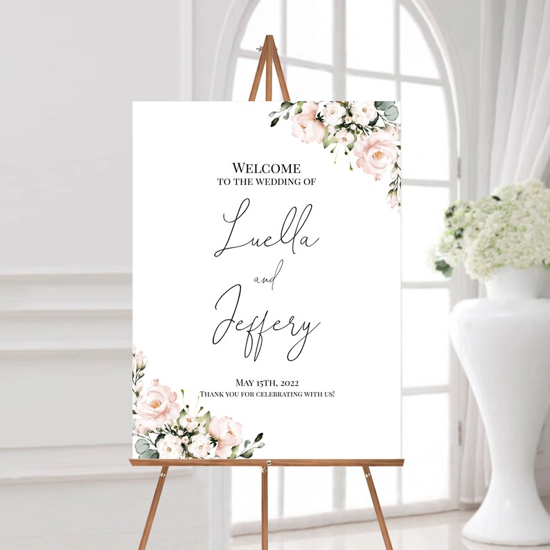 Puede incluir: Un cartel de bienvenida de boda blanco con un dise&ntilde;o floral en la esquina. El cartel dice "Bienvenido a la boda de Luella y Jeffery 15 de mayo de 2022 &iexcl;Gracias por celebrar con nosotros!"