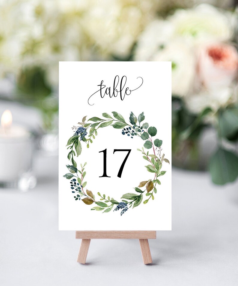 May include: White table number card with a green and blue floral wreath and the number 17 in the center. The card says "table" at the top and is displayed on a small wooden easel.