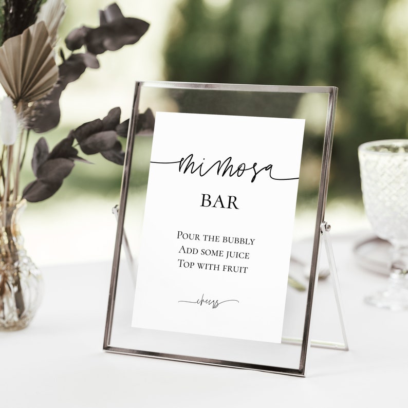 Pu&ograve; includere: Una cornice di vetro trasparente contiene una scheda bianca con testo nero: "Mimosa Bar". La scheda elenca le istruzioni: "Versare le bollicine, aggiungere un po' di succo, guarnire con frutta". La cornice &egrave; su una superficie bianca, con verde sullo sfondo.