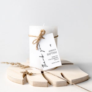 May include: A white pillar candle tied with twine, featuring a tag that reads "Adrian's Baptism" and "Thank you for celebrating with us." The candle rests on a wooden decorative piece. The text "thank you" is also written on the tag.
