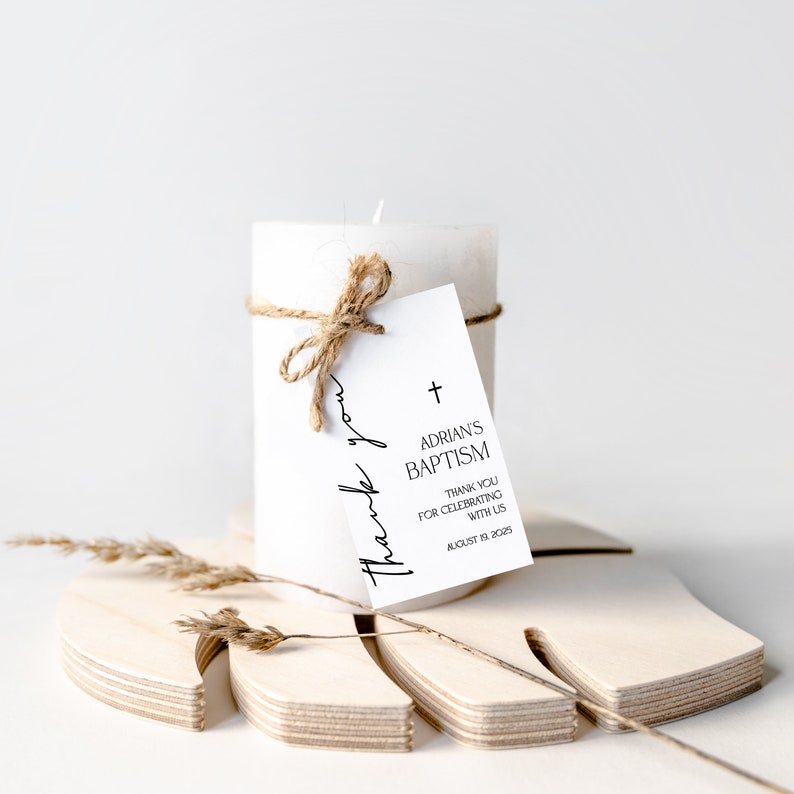 May include: A white pillar candle tied with twine, featuring a tag that reads "Adrian's Baptism" and "Thank you for celebrating with us." The candle rests on a wooden decorative piece. The text "thank you" is also written on the tag.