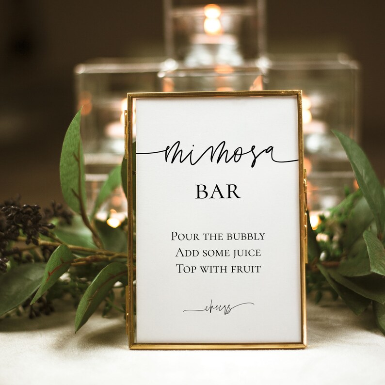 Pu&ograve; includere: Un cartello bianco in una cornice dorata con le parole "Mimosa Bar" in elegante scrittura nera. Sotto, le istruzioni recitano "Versare le bollicine, Aggiungere un po' di succo, Guarnire con frutta". La parola "cheers" &egrave; in basso.