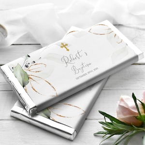 Puede incluir: Dos barras de chocolate envueltas en papel plateado con un fondo blanco con una cruz dorada, hojas verdes y blancas y purpurina dorada. El texto en la barra dice "Robert's Baptism September 28th, 2020".