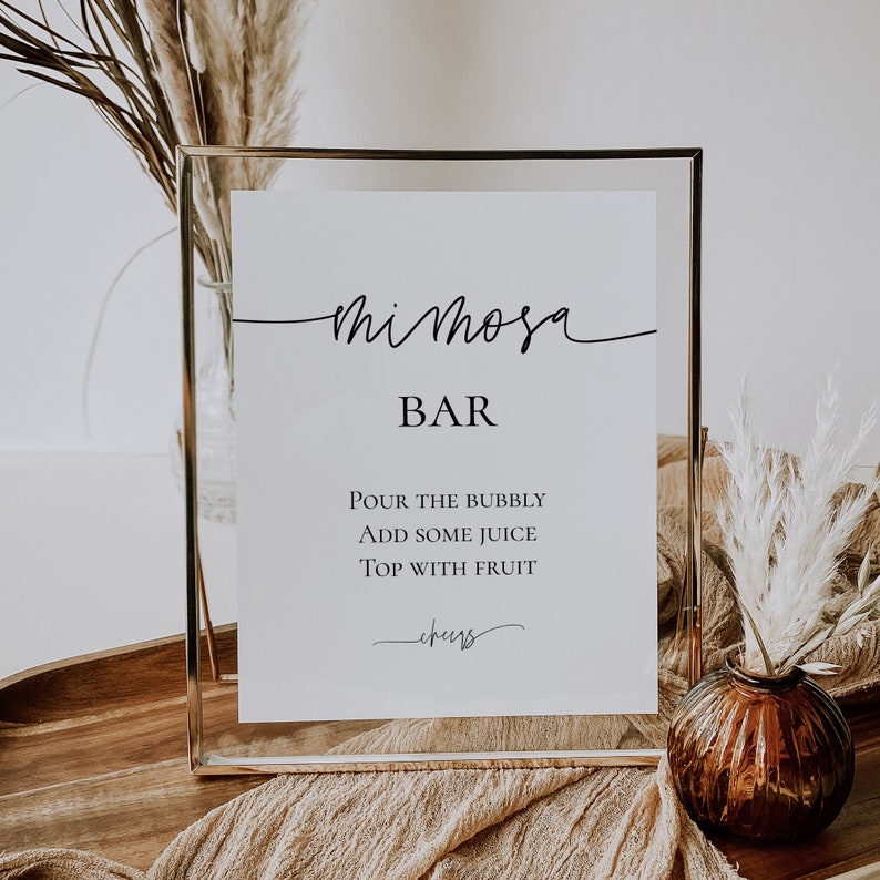 Pu&ograve; includere: Un cartello per il Mimosa Bar in una cornice dorata. Il cartello bianco presenta la scritta nera "Mimosa Bar" e le istruzioni: "Versare le bollicine, aggiungere un po' di succo, guarnire con la frutta". Un vaso decorativo e fiori secchi sono sullo sfondo.