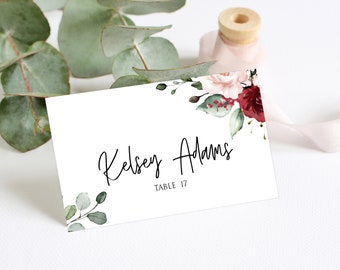 May include: White wedding place card with a floral design in shades of pink and burgundy. The card has the name "Kelsey Adams" and the text "Table 17" printed in black.