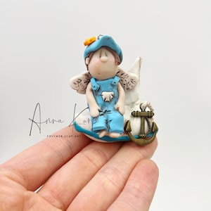 Puede incluir: Una pequeña figurita azul y blanca de un hada de los dientes sentada en una nube blanca con un ancla dorada. El hada lleva un sombrero azul, un mono y alas. El hada tiene el pelo castaño y una cara marrón claro.