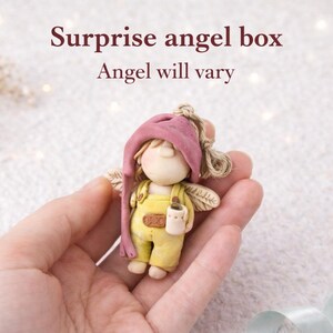 Könnte beinhalten: Handgefertigte Engelsfigur mit rosa Mütze und gelbem Overall, die einen kleinen Becher hält. Der Engel hat kleine Flügel und wird in einer Hand gehalten. Der Text "Surprise angel box" und "Angel will vary" steht darüber.