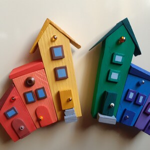 May include: A rainbow-colored set of miniature wooden houses with blue windows and doors. The houses are arranged in a row, with the tallest house in the center. The houses are painted in shades of red, orange, yellow, green, blue, and purple.