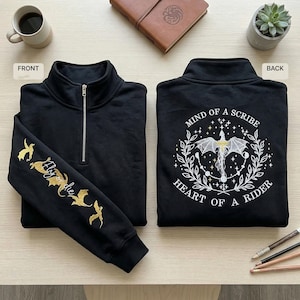 May include: Black quarter-zip sweatshirt with gold embroidery. The front features a zipper and the sleeve has the text "Fly the" with bat designs. The back has a dragon design with the text "Mind of a Scribe, Heart of a Rider."